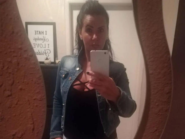 Lenore-hot taking a selfie, wearing a denim jacket over black top with criss-cross straps, holding a white phone, mirror reflection, brown hair in a messy ponytail, serious expression.