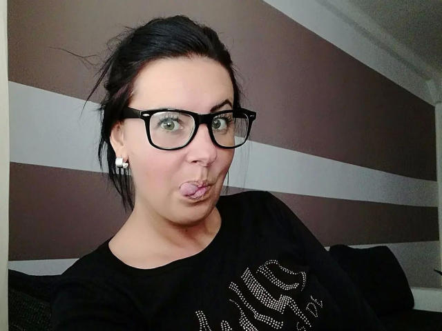 Lenore-hot with black glasses, puffy pink tongue out, black shirt, and earrings, has dark hair in a messy bun, sitting, looking at camera, in a selfie.
