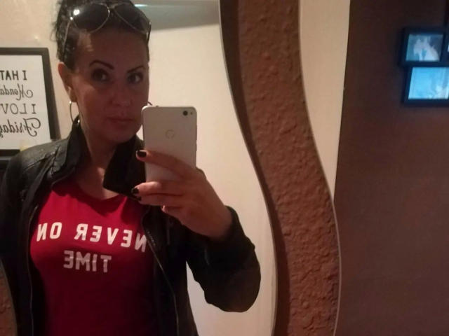 Lenore-hot with dark skin, wearing a red shirt with "NO REASON" and black jacket, holding a phone, taking a mirror selfie, eyewear on head, in a dimly lit room.