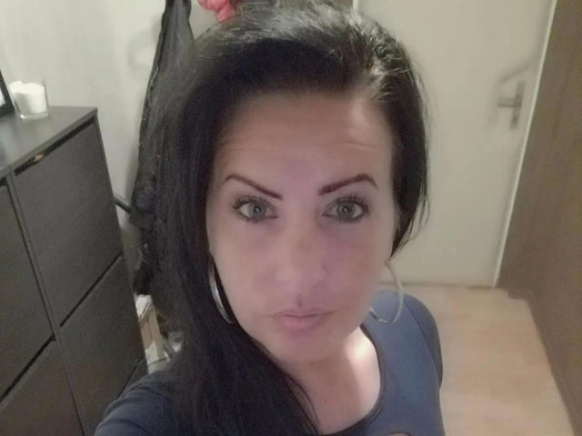 Lenore-hot has dark hair, green eyes, and a neutral expression. She's wearing a navy shirt, with her tits slightly visible under the shirt. She's looking at the camera.