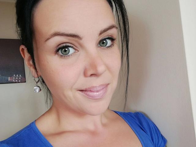 XxX-Melanie with green eyes, black hair in a ponytail, wearing a blue shirt, has shiny pink lips, and small, round, silver earrings, smiling slightly at the camera.