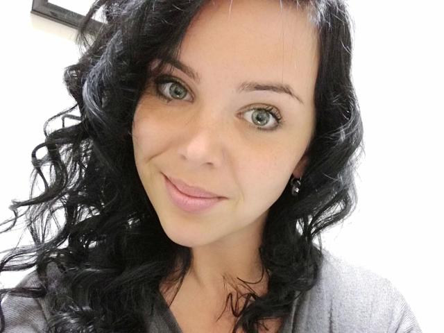 XxX-Melanie has curly black hair, green eyes, and a cute smile with pink lips. She's wearing a gray shirt and has small, subtle earrings.