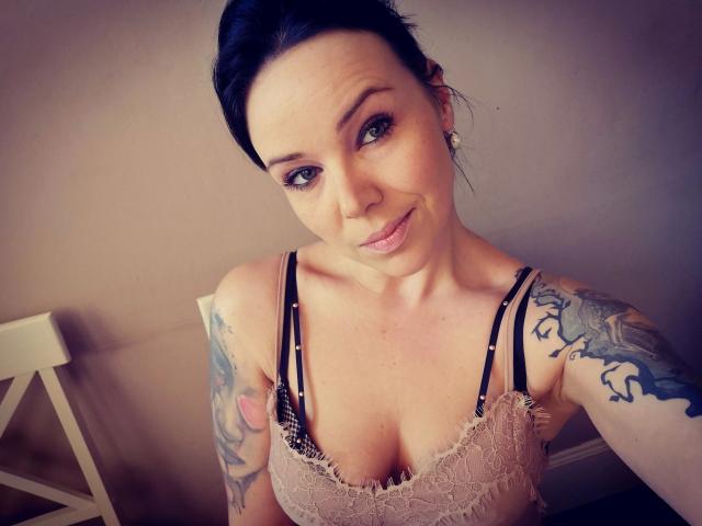 XxX-Melanie with dark hair, smiling, wearing lace bra, showing tattoos on right arm, small earrings, pale skin, and visible cleavage. She's looking up at the camera.