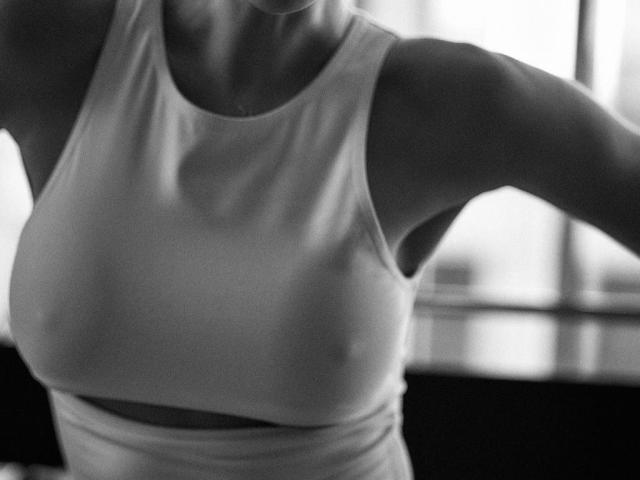 I seni di MarieAngel sono visibili attraverso una canotta bianca aderente, che mostra dei capezzoli appena accennati. La foto in bianco e nero enfatizza la parte superiore del suo corpo e le sue braccia muscolose..