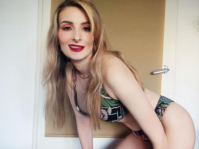 HeisseManuelle-hot with long blonde hair, red lipstick, and patterned bikini, leaning forward, showing her tits and ass, smiling, standing in front of a door.