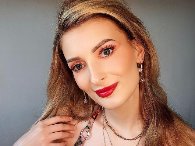 HeisseManuelle-hot ha lunghi capelli biondi con una mecca scura, rossetto rosso e eyeliner arancione. Indossa orecchini e una collana, e ha un sorriso sul viso..