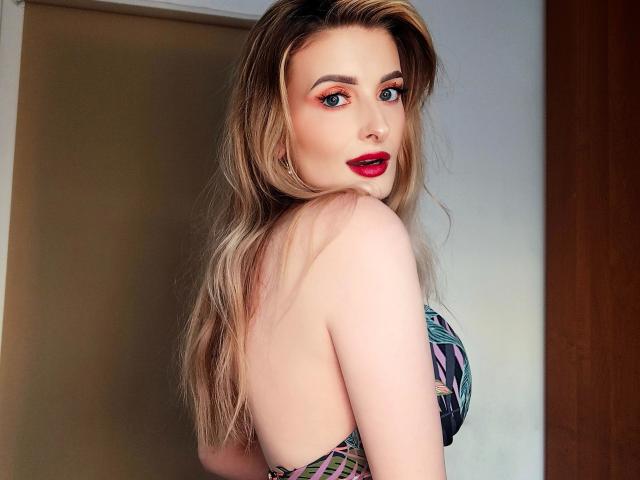 HeisseManuelle-hot with long blonde hair, red lipstick, and blue eyeshadow, looking over her shoulder, showing her back and patterned top, with her tits slightly visible.