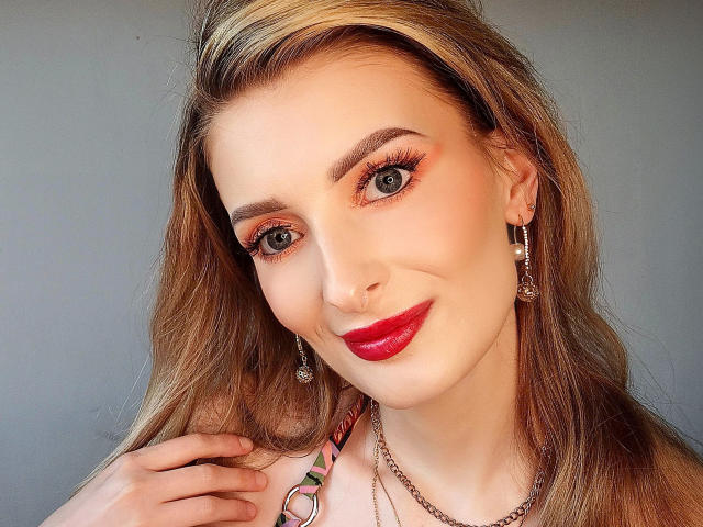 HeisseManuelle-hot has blonde hair, red lipstick, and orange eye makeup. She's smiling with her hand near her neck, wearing earrings and a necklace. Her tits are not visible.