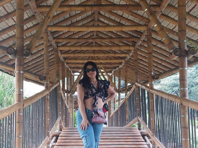 LauraRichis stands on wooden stairs, wearing a black floral top, blue jeans, and sunglasses, with a pink purse, under a bamboo-roofed structure. Her tits are slightly visible.