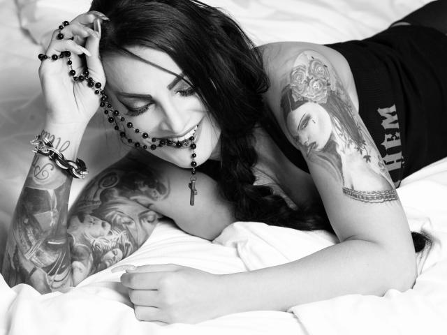 Sandra Sturm, attraente e con tatuaggi, sdraiata sul letto, indossa un top nero, ha un rosario in bocca, sorride, ha tatuaggi sulle braccia, una treccia, capelli neri, un letto bianco, un braccialetto nero, e appare sexy..