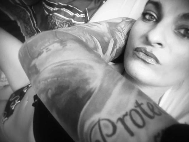 Black-and-white selfie of SandraSturm-hot with tattoos covering her right arm, showing "Prote" in cursive. She has dark lipstick and is lying down, partially visible.