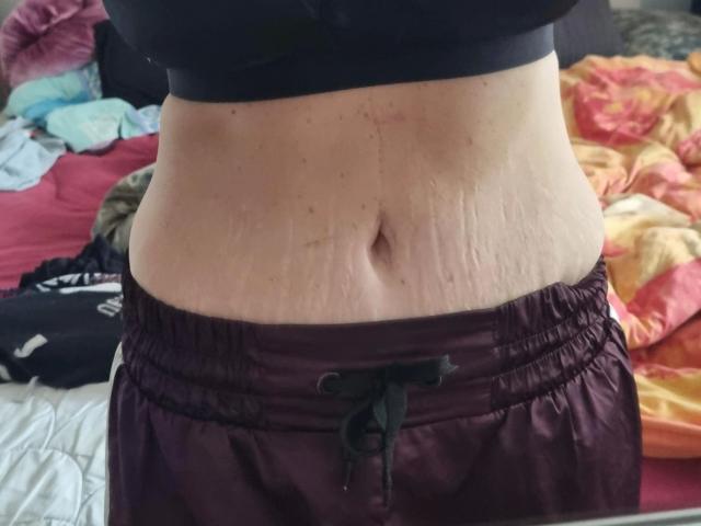 Wetgerman-hot's midsection with visible stretch marks, wearing dark maroon, shiny, loose-fitting shorts, black sports bra, and a slightly protruding belly button.