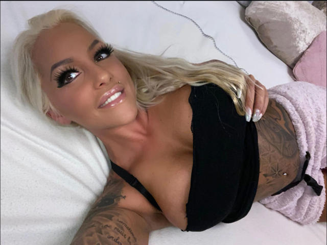 CocoSky-hot with blonde hair, large tits, tattoos, black top, pink pants, smiling, lying on bed, white sheets, long eyelashes, manicured nails, tan skin, sexy, attractive.