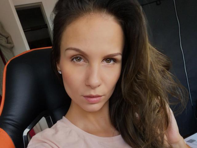 MaryWet-hot has light brown skin, green eyes, and long brown hair, wearing a pink shirt, sitting in a black and orange chair, looking straight at the camera.