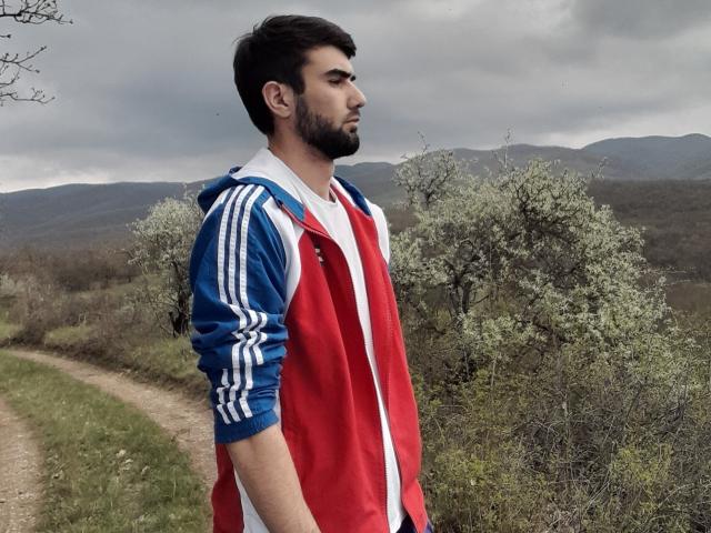 AliskerHot has a red and blue Adidas jacket, white shirt, and black pants, standing on a dirt path with trees and mountains in the cloudy background.