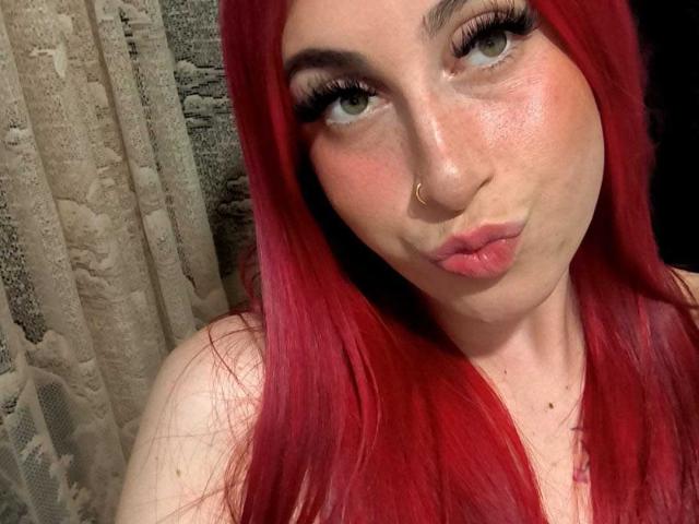 SammyViolet with red hair, pouty lips, green eyes, and a nose ring, giving a cute, slightly annoyed look. Boobs are not visible. She's wearing long, straight hair.