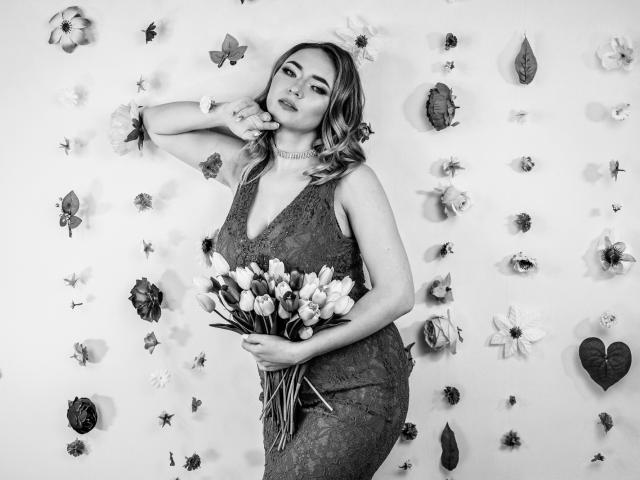 SallyVerda stands with one hand near her mouth, wearing a lace dress, holding a bouquet of tulips, and has her tits slightly visible, in a black-and-white photo.