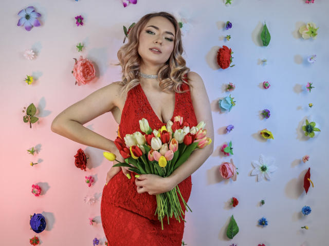 SallyVerda with blonde curls, wearing a red lace dress, holding a bouquet of yellow and red tulips, showing her tits, against a wall of colorful flower decorations.