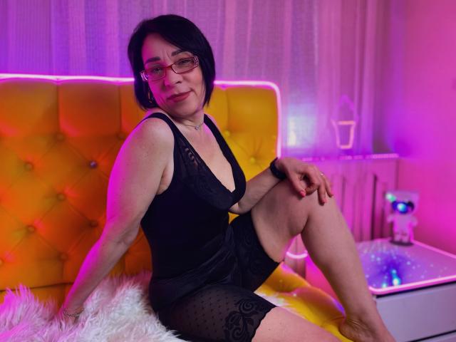 SandraHormas, avec ses cheveux noirs courts, ses lunettes et sa robe noire en dentelle, assise sur une tête de lit jaune capitonnée, les jambes croisées, avec des lumières roses et violettes, et un coussin blanc en fausse fourrure..
