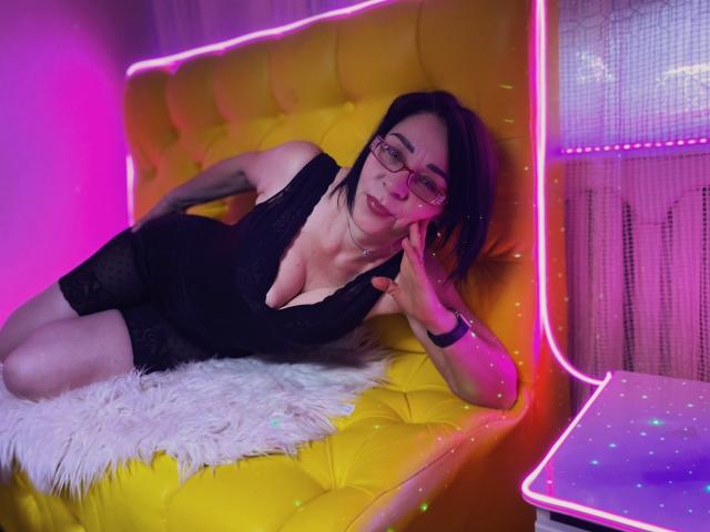 SandraHormas, avec des lunettes, une robe noire en dentelle et une poitrine de taille moyenne, est assise sur un canapé jaune capitonné, avec des néons roses, de la fourrure blanche et une table violette lumineuse..