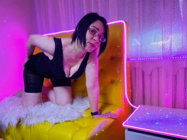 SandraHormas, mit schwartem Bob und Brille, kniet auf einem gelben, mit Noppen versehenen Sofa, trägt schwarze Dessous, zeigt große Brüste, und unter ihr liegt ein flauschiger, weißer Teppich, beleuchtet von neonrosa und violetten Lichtern..