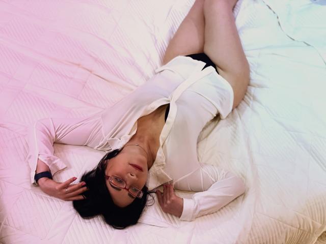 SandraHormas è sdraiata su un letto, indossa una camicia bianca sbottonata che lascia intravedere delle mutandine nere, le gambe sono unite, porta occhiali da vista neri e la bocca è leggermente aperta..