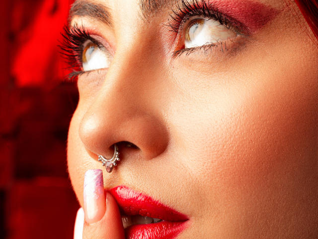 IsabellaRobertz' Gesicht mit rotem Make-up, einem Nasenring und rotem Lippenstift, blickt nach oben mit braunen Augen, berührt ihre Lippe mit einem violett lackierten Finger, vor einem roten Hintergrund..