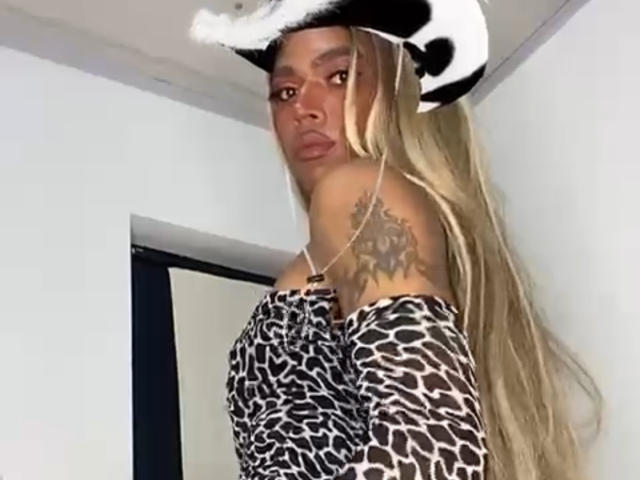 SarhaBlackTs with long blonde hair, wearing a cow hat and leopard print off-shoulder top, showing a sun tattoo on her right shoulder, looking at the camera.