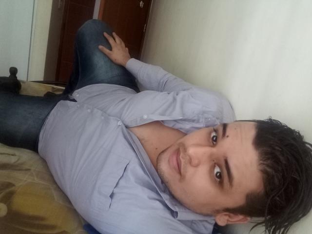 Bigchef is lying on a bed, wearing a blue shirt, and blue jeans, with a hand on his knee, looking at the camera, and showing a slight smile.