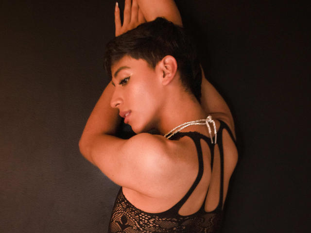 Anddyys with short black hair, arms raised, wearing a black lace top with strappy back, showing off a pearl necklace, against a dark background, looking to the side.