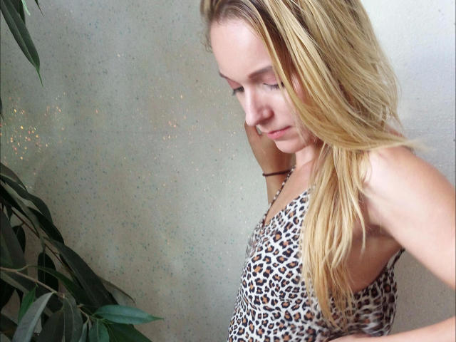 Lolicoon - com cabelo loiro, vestindo uma blusa com estampa de leopardo, olhando para baixo, com o braço levantado, seios pequenos, bumbum discreto, em pé ao lado de uma planta verde com folhas..