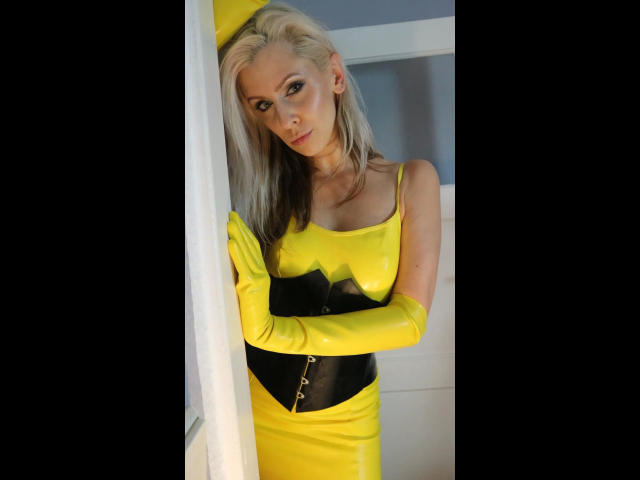 Lady Sybella - atraente, com cabelo loiro, vestindo um vestido de látex amarelo, um painel preto no peito e luvas amarelas, apoiada em uma parede branca, olhando para a câmera com uma expressão sedutora..