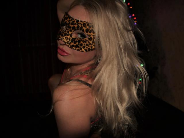 JanePussyCam-hot with long blonde hair, wearing a leopard mask, pink lipstick, and a red bra, looking over her shoulder in a dimly lit room with colorful lights.
