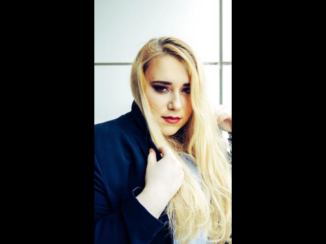 HaileyBLONDE-hot with long blonde hair, red lipstick, and dark eyeliner, wearing a navy blue jacket, holding her hair, looking slightly to the side, with a subtle smile.