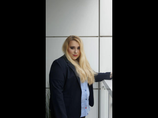 HaileyBLONDE-hot with long blonde hair, wearing a dark blazer over a light blue button-up shirt, standing by a metal railing, looking slightly down with a subtle smile.