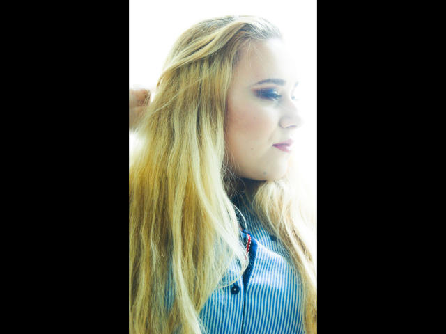 HaileyBLONDE-hot with long blonde hair, blue striped shirt, and buttoned chest, looking to the side with a slight smile, right hand in hair. Bright, overexposed lighting.