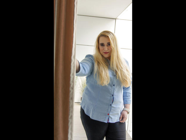 HaileyBLONDE-hot with long blonde hair, wearing a blue button-up shirt and black pants, standing in a doorway, extending her right arm, looking confident.