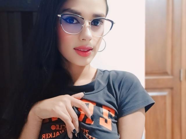 AnaisHilton with long black hair, blue eyeshadow, glasses, red lipstick, gray t-shirt, and black nail polish, touching her chest with fingers. Wooden door in background.