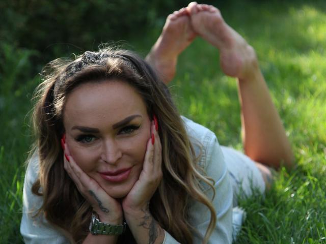 PenelopeLopez-hot lying on grass, face in hands, smiling, green eyes, wavy brown hair, red nails, white shirt, tattoo on hand, green watch, feet up, sunlight.