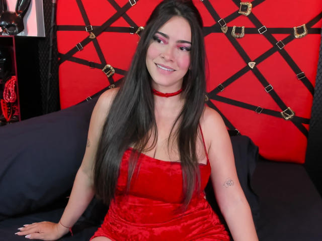 ChristineBray with long black hair, red velvet dress, red choker, smiling, pink eyeshadow, black tattoo on right arm, sitting on black bed, red and black wall behind.