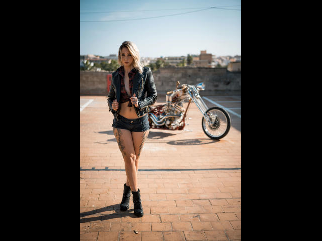 MimiSweet-hot with tattoos, wearing a denim jacket, black shorts, and boots, standing on a rooftop with a chopper bike in the background. Her ass and tits are visible.