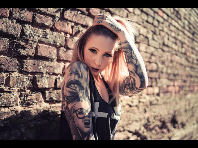 MimiSweet-hot with tattoos, pulling up her blonde hair, wearing a black tank top, standing against a brick wall, looking at the camera with dark lipstick.