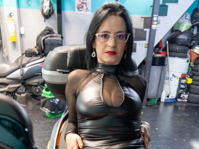 AlanaMoneth with black hair, glasses, red lipstick, and a shiny black leather dress with a cutout, sitting in a car seat, her tits prominently visible.