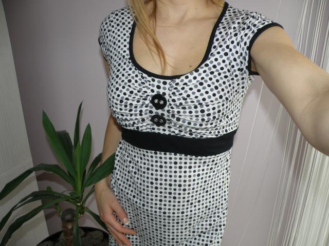 NNataszka-hot wearing a white dress with black polka dots, black buttons, and a black waistband. Her hand is near her crotch, and a green plant is visible.