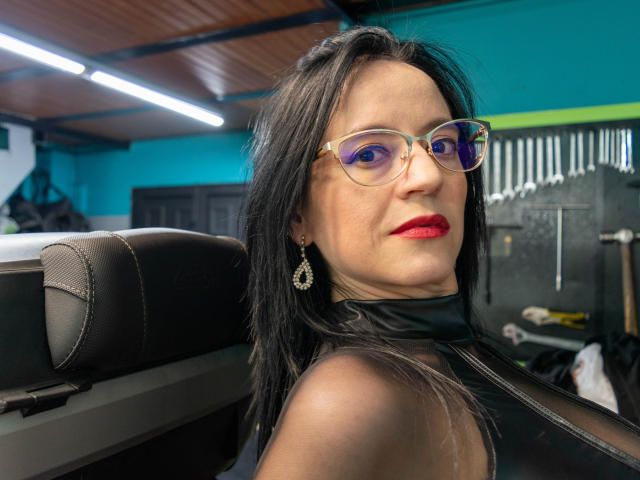 AlanaMoneth with red lipstick, black hair, clear glasses, and teardrop earrings, wearing a black leather top, sitting in a car with brown leather seats.