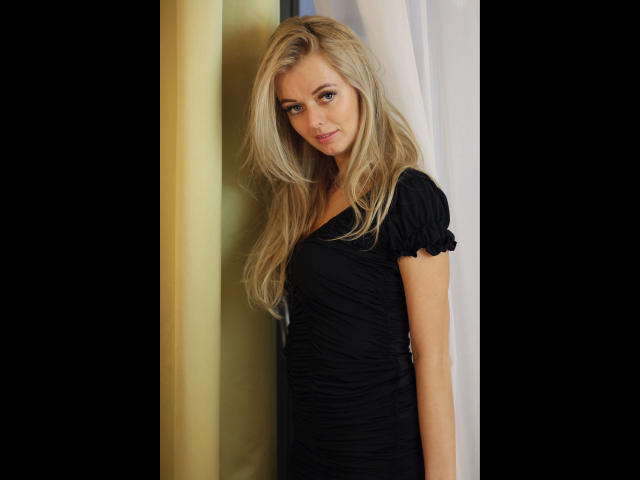 Sensual Valerie - linda, com longos cabelos loiros, vestido preto franzido e um sorriso adorável, em pé em frente a uma cortina amarela e branca, parecendo sexy com seus olhos azuis e um leve sorriso..