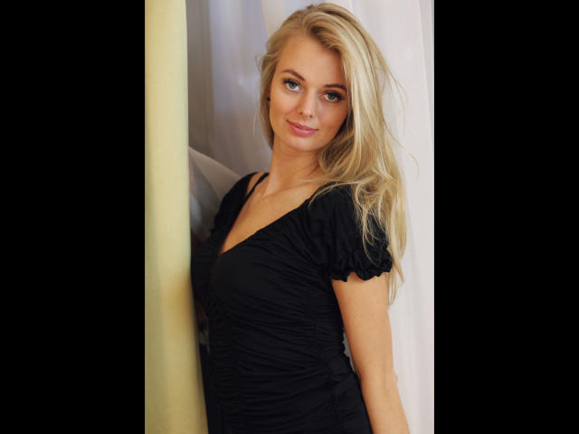 Sensualvalerie: atractiva, con cabello rubio, ojos azules y un vestido negro ajustado, de pie junto a una pared amarilla, sonriendo sutilmente, mostrando un pequeño atisbo de su trasero..
