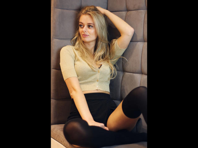 Sensualvalerie-hot has blonde hair, small tits, wears a yellow crop top and black skirt, black thigh-highs, sitting with one hand in her hair, and a relaxed, sexy smile.