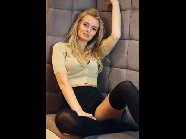 Sensual Valerie - atraente, com cabelo loiro, vestindo uma cropped amarela, saia preta e meias pretas até a coxa, sentada com um braço levantado, mostrando seus pequenos seios e vulva..