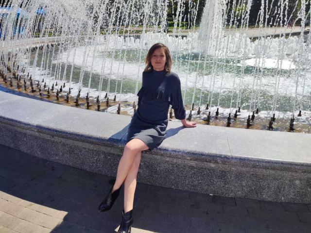 AngelaRoxyx sitzt auf einer Steinsitzbank, trägt ein dunkelblaues Strickkleid und schwarze Stiefeletten, und hinter ihr befindet sich eine Springbrunnen. Ihre Beine sind gekreuzt, und sie lächelt..